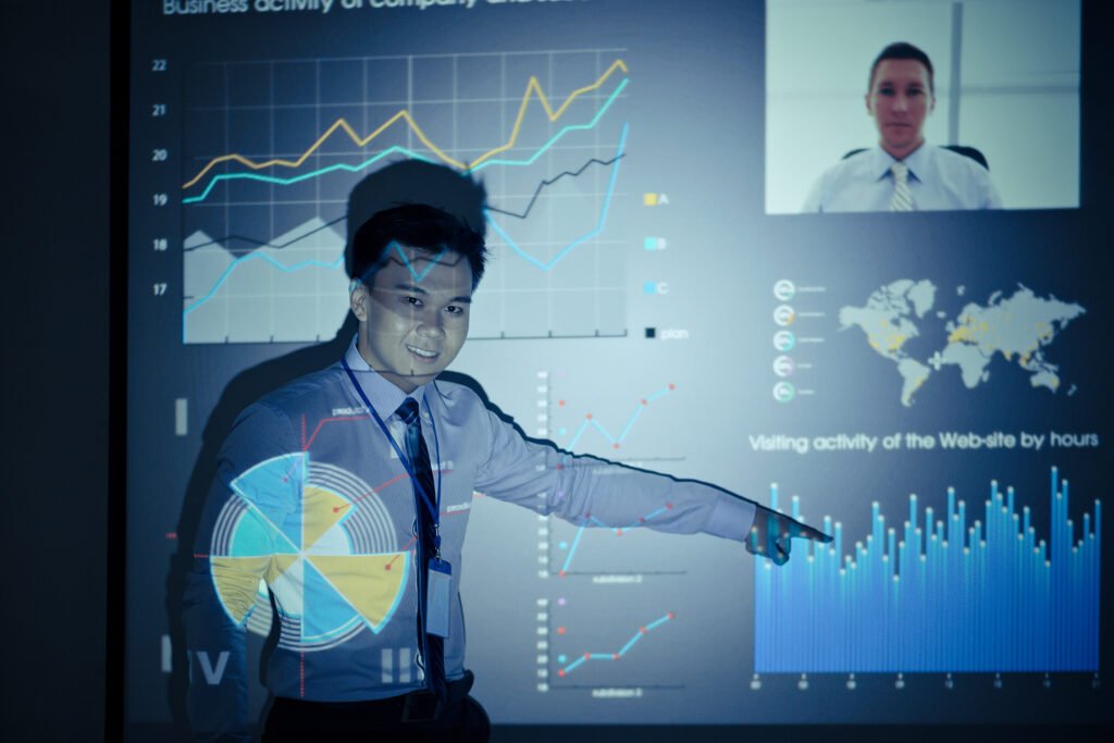 a Person in a Shirt and Tie Gives a Presentation with Charts and Graphs Projected on a Screen, Discussing Business Metrics and Aspects of It Project Management, While a Video Call Participant is Visible.