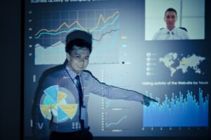 a Person in a Shirt and Tie Gives a Presentation with Charts and Graphs Projected on a Screen, Discussing Business Metrics and Aspects of It Project Management, While a Video Call Participant is Visible.