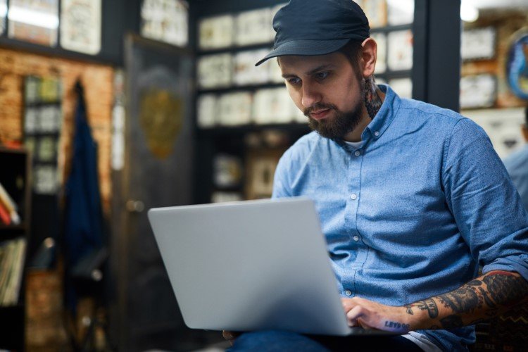 a Man with Tattoos and a Beard, Wearing a Blue Shirt and Black Cap, is Sitting and Working on a Laptop in a Room with Framed Artwork on the Walls, Likely Collaborating with It Teams.