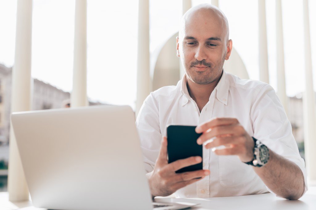 a Man Sitting at a Table with a Laptop, Immersed in Remote It Project Management, Glances at His Phone.