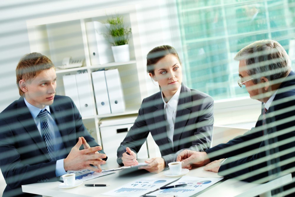 Three Consulting Professionals in Business Attire Are Engaged in a Meeting Around a Table with Documents, Charts, and Coffee Cups, Viewed Through Window Blinds.