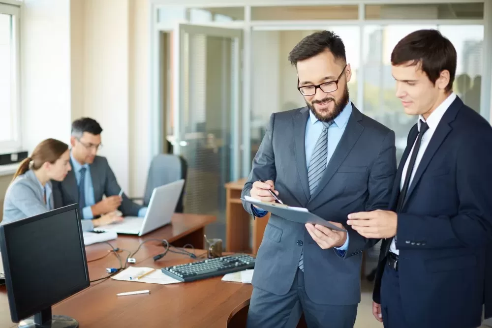 Two Men in Suits Exploring a Document, with Colleagues Working in the Background. It Consulting Practices