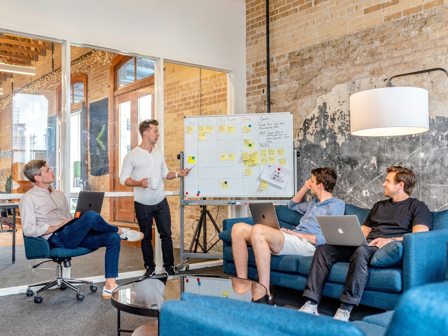 Four people in a casual office setting are having a meeting. One person, possibly from IT Consultants, stands by a whiteboard with notes on top focus areas, while the others sit on couches with laptops, attentively listening.