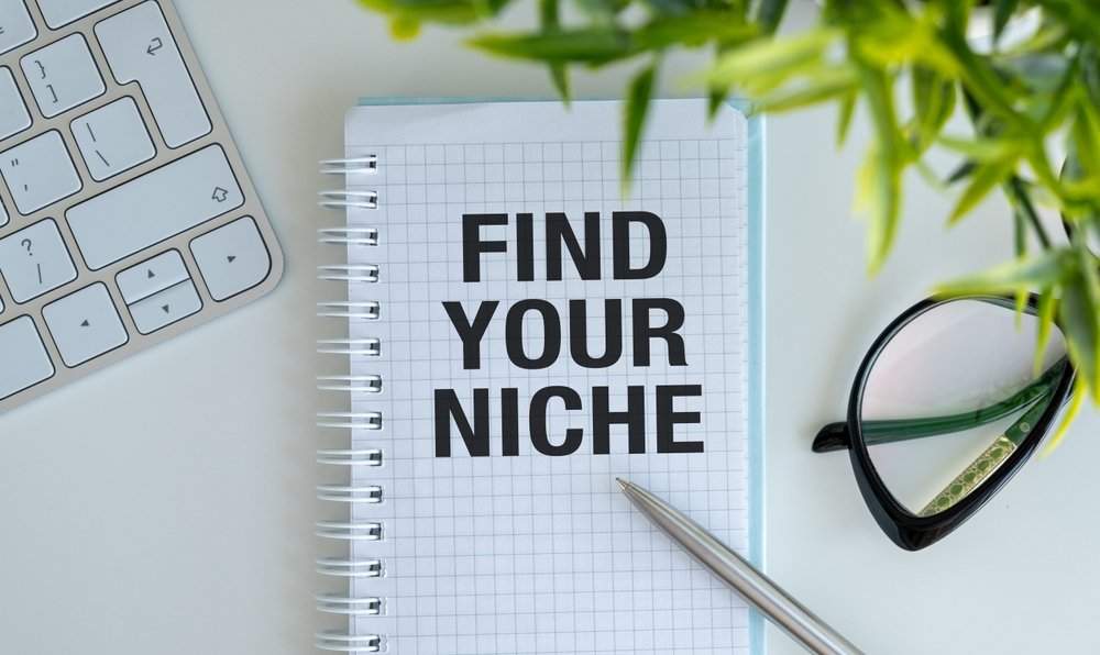 Notebook with "find Your Niche" Text Beside a Keyboard, Glasses, and a Plant—perfect for Jotting Down Ideas on Niche Expertise in It Consulting.