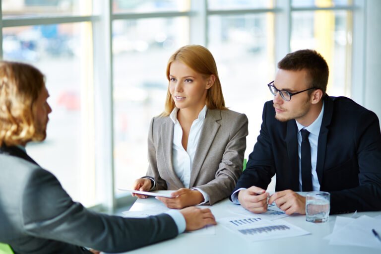 Three professionals in business attire sit at a table in an office, deeply engaged in a "Business Now" discussion. Documents and a glass of water are spread before them, as they strategize on IT consulting solutions to boost your business efficiently.