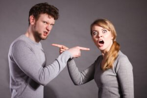 a Man and Woman in Gray Shirts Furiously Point at Each Other, Standing Against a Gray Background, As if Caught in an Online Debate About Politics.