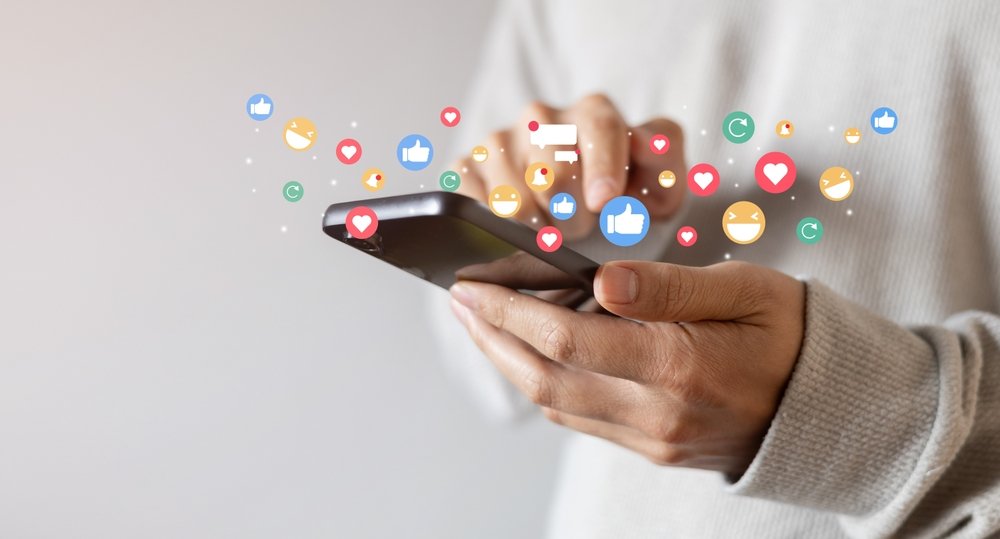 A person uses a smartphone with social media reaction icons, such as thumbs up, hearts, and emojis, appearing above the screen.