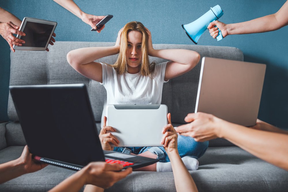 A woman sits on a couch holding her head, surrounded by multiple hands offering electronic devices and a megaphone, appearing overwhelmed by constant news updates and the demands of American News Media reporting.