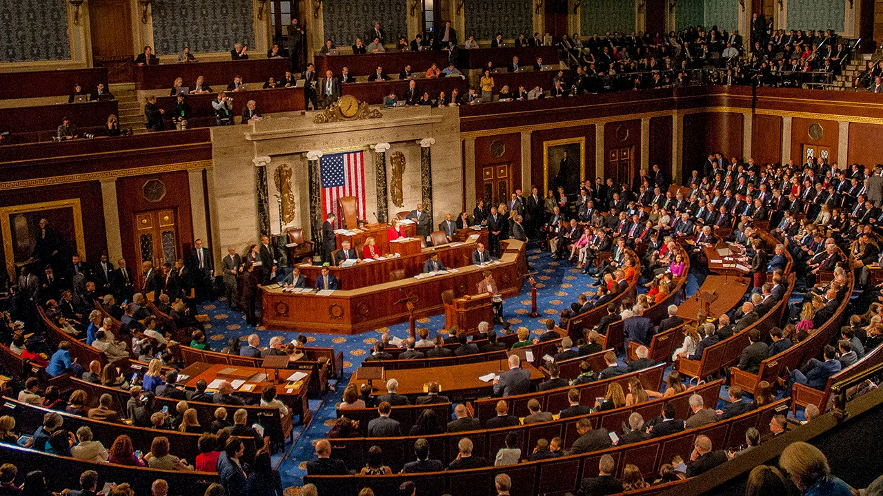 A wide view of the U.S. House of Representatives chamber filled with people during a formal Congress session, with the American flag displayed at the front, highlighting their mission of governing the nation.