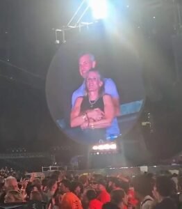 a Couple is Shown Hugging on a Large Circular Kiss Cam Screen Above a Crowded Concert Audience Inside an Indoor Arena.