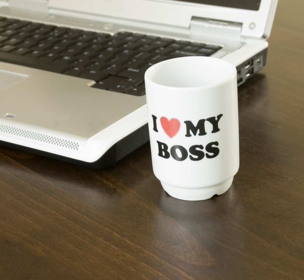 A white mug with "I ♥ MY BOSS" printed on it is placed on a wooden desk next to an open laptop—a playful touch for anyone focused on career advancement.