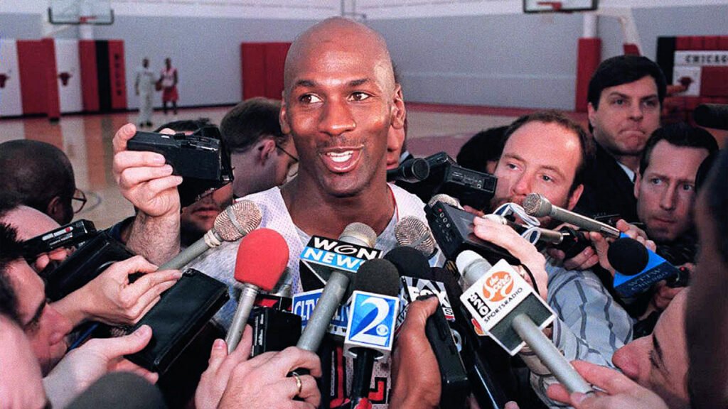 a Basketball Player Smiles While Surrounded by Reporters and Microphones During a Media Interview in a Gym, Discussing How Teamwork on the Court Can Impact the Bottom Line in Business.