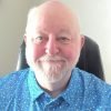 An older man with a white beard and mustache is sitting on a black chair, wearing a blue patterned shirt, and smiling at the camera. An older man with a white beard and mustache is sitting on a black chair, wearing a blue patterned shirt, and smiling at the camera.